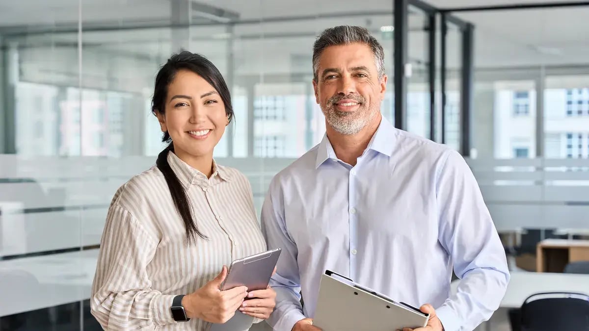 Two smiling colleagues holding a tablet and a clipboard in a bright office